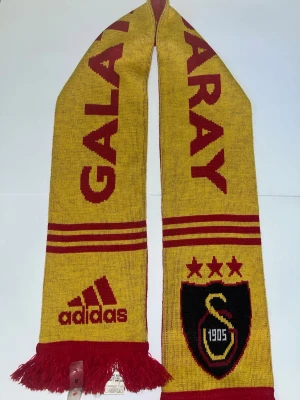 Galatasaray halsduk Adidas gul/röd - Snygg halsduk från Adidas med Galatasaray-tryck i gult och rött. Ena sidan är gul med röda detaljer och den andra röd med gula detaljer. Halsduken har klubbens emblem, tre stjärnor och fransar i ändarna. Perfekt för att visa ditt stöd på matchen!