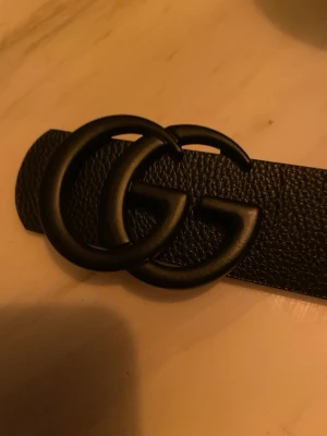 Svart skärp med GG-spänne från Gucci - Snyggt svart skärp från Gucci med det ikoniska dubbla GG-spännet i matt svart metall. Bältet är gjort i strukturerat skinn och har en klassisk, stilren design som passar till det mesta. Perfekt accessoar för att lyfta vilken outfit som helst.
