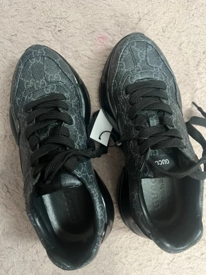 Svarta sneakers från Gucci - Snygga svarta sneakers från Gucci med GG-monogram över hela skon. Skorna har svarta snören, robust sula och är tillverkade i textil och läder. Perfekta för dig som vill ha en lyxig och trendig look.