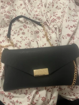 Svart kuvertväska från Valentino - Stilren svart kuvertväska från Valentino med guldfärgad kedjerem och elegant gulddetalj framtill där märkesnamnet syns. Väskan har en klassisk rektangulär form och är tillverkad i ett strukturerat material som ger en lyxig känsla. Perfekt accessoar för att lyfta din outfit.