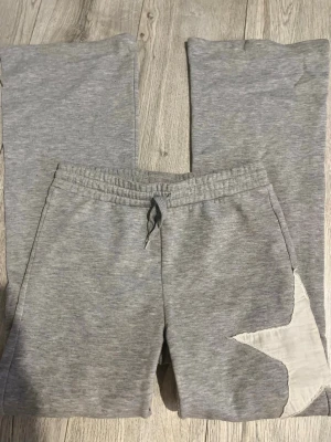 Grå mjukisbyxor med stjärndetalj - Säljer ett par grå mjukisbyxor med resår i midjan och dragsko. Byxorna har en vit stjärnformad detalj på ena benet och raka ben. Perfekta för chill dagar och riktigt sköna att ha på sig. Materialet känns mjukt och stretchigt.