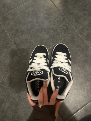Svarta Adidas sneakers med vita ränder - Svarta Adidas sneakers med klassiska vita ränder på sidorna och vit sula. Skorna har breda vita skosnören och Adidas logga på plösen. Ovandelen verkar vara i mocka eller textil och insidan är vit. Perfekta för en sportig och stilren look.