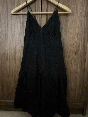 Svart blommig klänning med tunna band - Säljer en svart klänning med tunna axelband och v-ringning. Klänningen har blommiga broderier över hela tyget och en lätt utsvängd kjol. Perfekt för dig som gillar en stilren men ändå detaljrik look.