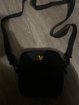 Svart axelväska från Lyle & Scott - Snygg svart axelväska från Lyle & Scott med justerbar axelrem och guldfärgad logga framtill. Väskan har en fyrkantig form och är tillverkad i slitstarkt tygmaterial. Perfekt för att bära det viktigaste och ger en clean look till din outfit.jätte fin och oanvänd!