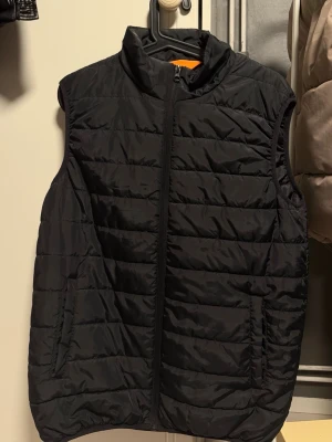 Svart quiltad dunväst - Stilren svart dunväst med quiltat mönster och hög krage. Västen har dragkedja framtill och två sidofickor. Perfekt för lager-på-lager och håller dig varm under kyliga dagar. Materialet är lätt och smidigt, vilket gör den enkel att bära med sig.