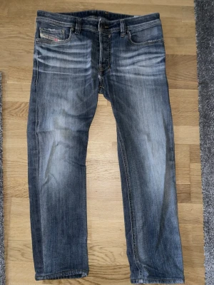 Blå jeans från Diesel med slitningar - Säljer ett par snygga blå jeans från Diesel med coola slitningar och tvättade detaljer. Klassisk femficksmodell med logga på myntfickan och kontrastsömmar. Jeansen har rak passform och är tillverkade i bomull. Och har lite små skador