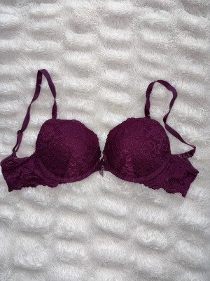 Vinröd/lila push-up bh med spets - Snygg vinröd/lila push-up bh med vadderade kupor och vacker spetsdetalj över hela bh:n. Justerbara axelband och hakknäppning bak. Liten dekorativ rosett framtill. Perfekt för dig som vill ha både stöd och en feminin look. Storlek 75A och knappt använd, Från lindex tror jag