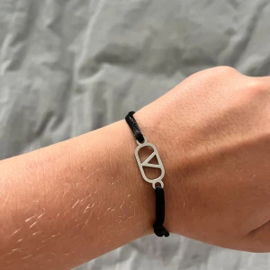 Svart armband med silverdetalj - Stilrent svart armband med en cool silverfärgad metallberlock i form av en geometrisk design. Justerbart band som passar de flesta handleder. Perfekt accessoar för att ge din outfit en modern touch.