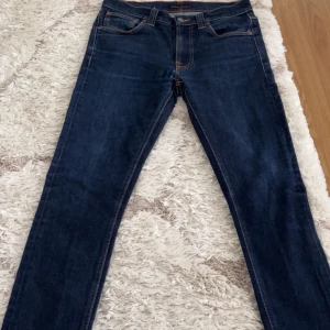 Mörkblå jeans från Nudie Jeans - Snygga mörkblå jeans från Nudie Jeans med klassisk femficksdesign och kontrastsömmar. Jeansen har raka ben och normal passform, tillverkade i slitstarkt denimtyg av bomull. Bakfickorna har Nudie Jeans signaturbrodyr i orange.