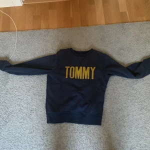 Mörkblå Tommy Hilfiger sweatshirt - Mörkblå sweatshirt från Tommy Hilfiger med stort gult tryck 'TOMMY' på bröstet. Klassisk rund halsringning och långa ärmar. Skön och stilren tröja i bomull, perfekt för en chill och snygg look.