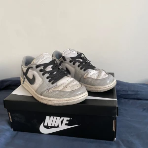 Nike Air Jordan 1 Low  - Säljer ett par Nike Air Jordan 1 Low i grått och vitt med svarta detaljer och snörning. Skorna har klassisk rund tå, perforerad tåbox och svart swoosh på sidan. Perfekta för dig som gillar streetwear och vill ha en ikonisk sneaker i garderoben.