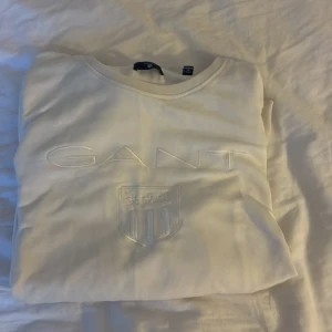 Vit sweatshirt från GANT - Säljer en stilren vit sweatshirt från GANT med broderad logga och emblem framtill. Tröjan har rund halsringning och långa ärmar, perfekt för en clean och klassisk look. Materialet är mjukt och skönt, passar till många outfits.