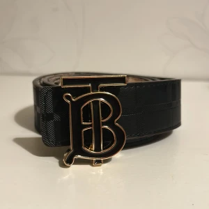 Burberry svart och rutig skärp i skinn - Snyggt Burberry-bälte i svart skinn med klassiskt rutmönster och präglad logga. Bältet har en stor guldfärgad spänne med TB-monogram och insidan har den ikoniska beigea Burberry-rutan med röda och svarta detaljer. Perfekt accessoar för att lyfta din outfit. Storlek 120