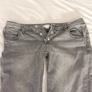 Grå jeans  - Säljer ett par gråa jeans från Gina Young i storlek 158, GRÅ LOW WAIST FLARE BUTTON JEANS,Flarejeans från Gina Tricot Young. Midjan är låg och gylfen består av knappar. Midjan är justerbar så att jeansen ska sitta så bra och bekvämt som möjligt. Benen har en smal passform och är utsvängda nedtill. Skriv om ni har mer frågor🥰  