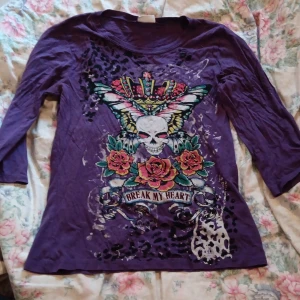 Lila topp med döskalletryck från Avanti - Cool lila topp från Avanti med trekvartsärm och stort färgglatt tryck framtill med döskalle, rosor, fjäril och krona. Texten 'Break My Heart' och leopardmönsterdetaljer gör den extra edgy. Tillverkad i mjuk bomull, perfekt för dig som gillar statement-plagg.