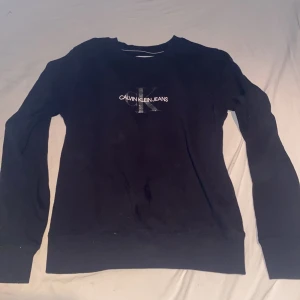 Svart Calvin Klein Jeans tröja - Svart långärmad tröja från Calvin Klein Jeans med klassisk logga i vitt och grått framtill. Tröjan har rund halsringning och är tillverkad i mjuk bomull, perfekt för en stilren och enkel look.