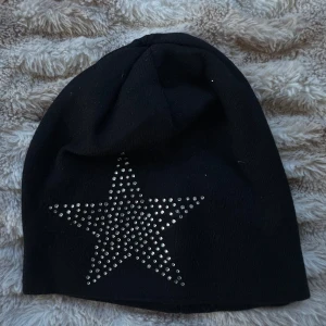 Yesstyle Svart mössa - Cool svart mössa med en stor stjärna framtill, dekorerad med silvriga nitar som ger extra bling. Mössan har en enkel, lite slouchy form och är tillverkad i mjukt material som passar perfekt för kyliga dagar. Perfekt accessoar för dig som gillar att sticka ut med detaljer.