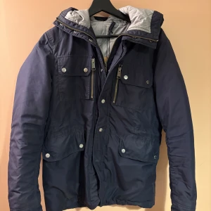 Fieldjacket från J.Lindeberg - Fieldjacket från J.Lindeberg i mörkblått med grått foder och huva. Jackan har flera fickor med knappar och dragkedjor framtill, samt hög krage och justerbar huva. Perfekt för kyliga dagar och har en stilren look. Skick 10/10 ☺️