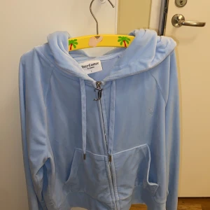 Ljusblå velour hoodie Juicy Couture - Ljusblå hoodie i mjuk velour från Juicy Couture med huva, snörning och dragkedja med J-hänge. Fickor framtill och glittrande Juicy-text i strass på ryggen. Perfekt för dig som vill ha en ikonisk och comfy look.