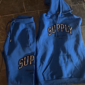 Blå dress, Tröja M, Byxor L - Blå hoodie från Supply med stor vit och svart text på bröstet. Tröjan har huva med snörning och en stor magficka. Perfekt för dig som gillar streetwear och vill ha en skön och avslappnad stil.