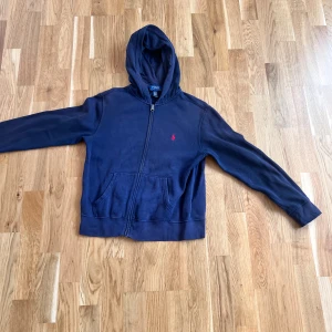 Marinblå hoodie från Polo Ralph Lauren - Snygg marinblå hoodie från Polo Ralph Lauren med dragkedja och huva. Tröjan har en röd broderad logga på bröstet, känguruficka framtill och är tillverkad i mjuk bomull. Perfekt för chill dagar eller när du vill ha en klassisk streetstyle-look.
