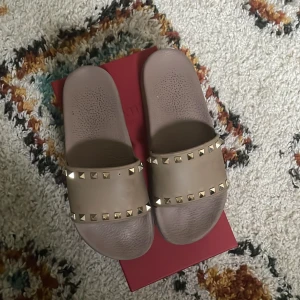 Valentino Rockstud sandaler beige strl 40 - Beiga Valentino Rockstud sandaler i PVC med ikoniska nitar i guld längs remmen. Platt sula och öppen tå ger en chill vibe. Perfekta för sommaren och lätta att matcha med allt. Kommer med originalkartong och extra nitar. En spik saknas.