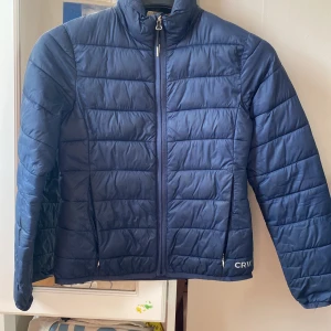 Marinblå pufferjacka från CRW - Säljer en marinblå pufferjacka från CRW med quiltad design och dragkedja framtill. Jackan har hög krage och två fickor med dragkedja. Perfekt för kyliga dagar och enkel att matcha med olika outfits.