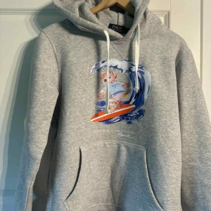 Grå hoodie Polo Bear Ralph Lauren - Grå hoodie från Polo Ralph Lauren med ikoniska Polo Bear-trycket där björnen surfar på en våg. Tröjan har huva med vita snören och en stor magficka. Mjuk bomullsblandning och långärmad modell, perfekt för chill dagar.