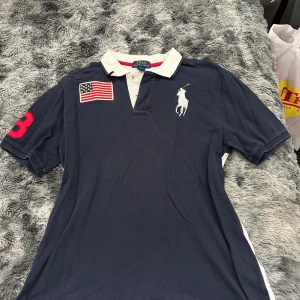 Mörkblå Polo Ralph Lauren t-shirt XL - Snygg mörkblå t-shirt från Polo Ralph Lauren med vit krage och knappar. Stor amerikansk flagga och röd siffra 3 på ärmen, samt klassisk vit Polo-logga på bröstet. Perfekt för dig som gillar sportig och preppy stil.