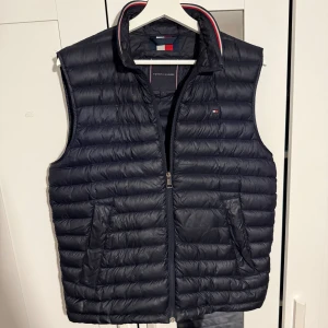 Mörkblå dunväst från Tommy Hilfiger - Snygg mörkblå dunväst från Tommy Hilfiger med TH Tech, quiltad design och dragkedja framtill. Västen har ståkrage med röd och vit detalj, två fickor och liten logga på bröstet. Perfekt för lager-på-lager och enkel att packa ner. Finns ett litet märke som syns på sista bilden men enligt mig tycker ja de knappt syns alls. Går ju absolut att laga med för den som vill de.