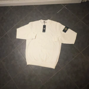 Vit långärmad tröja från Stone Island - Vit långärmad tröja från Stone Island med klassisk rund hals och ikonisk logotyp-patch på vänster ärm. Tröjan är i mjuk bomull och har en clean, stilren look som passar perfekt till jeans eller joggers.