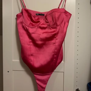Rosa satin body från Zara - Säljer en snygg rosa body från Zara i glansigt satinmaterial. Bodyn har tunna justerbara axelband och formade kupor för en cool och feminin look. Perfekt att styla med jeans eller kjol för en trendig outfit.