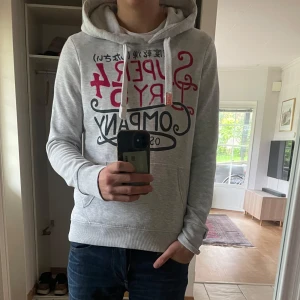 Grå vintage Superdry hoodie med tryck - Säljer en grå hoodie från Superdry med stort tryck i rött och svart på bröstet. Tröjan har huva med snörning, känguruficka och ribbade muddar. Perfekt för dig som gillar streetwear och vill ha en snygg och bekväm look.