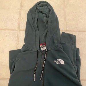 Mörkgrön hoodie från The North Face M - Säljer en mörkgrön hoodie från The North Face i storlek M. Tröjan har dragkedja, huva med snörning och logga på bröstet. Tillverkad i en mjuk blandning av bomull och polyester, perfekt för kyliga dagar.