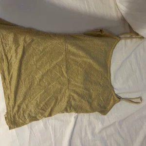 Glittrig guldig linnetopp - Snygg guldig linnetopp med smala axelband och glittrigt tyg. Toppen har en rak passform och är perfekt för dig som vill sticka ut med en metallic look. Materialet är mjukt och följsamt, vilket gör den bekväm att bära. Perfekt att styla med jeans eller kjol för en trendig outfit.