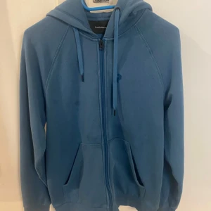 Blå hoodie från Peak Performance S - Snygg blå hoodie från Peak Performance med dragkedja och huva. Tröjan har snörning i halsen, två fickor fram och ett diskret logotryck på bröstet. Perfekt för chill dagar eller när du vill ha en sportig look. Storlek S