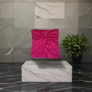 Rosa bandeau tubtopp med hjärtspänne - Snygg rosa bandeau tubtopp med ett glansigt tyg och ett dekorativt hjärtformat spänne med pärlor framtill. Toppen har draperad front och en liten volangdetalj som hänger ner. Perfekt för dig som vill sticka ut med en färgstark look.