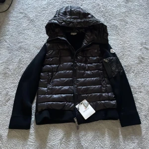 Moncler cardigan - Svart cardigan från Moncler med quiltad framsida och huva. Jackan har stickade ärmar och detaljer, dragkedja framtill och en liten ficka med dragkedja på ena ärmen. Klassisk Moncler-logga på ärmen och snygg, sportig look.