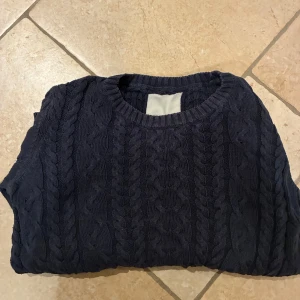 Mörkblå stickad - Mörkblå kabelstickad tröja från 157 med rund halsringning och ribbade muddar. Tröjan har ett klassiskt flätmönster och är långärmad, perfekt för kyligare dagar. Snygg och enkel att matcha med jeans.