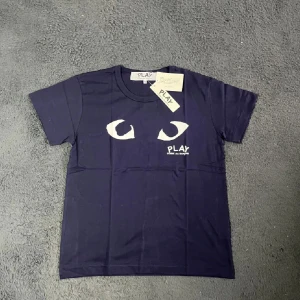 Mörkblå Comme des Garçons Play t-shirt - Snygg mörkblå t-shirt från Comme des Garçons Play med vit tryck av ögon på bröstet och logga. Klassisk rund hals och korta ärmar. Materialet är mjuk bomull som känns skönt mot huden. Perfekt för dig som gillar streetwear och vill sticka ut med en unik design.