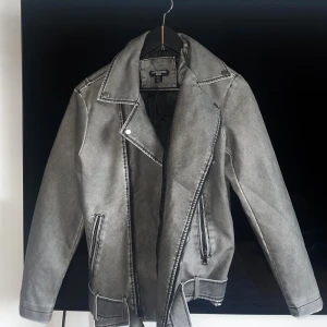 Grå bikerjacka från PLT (helt oanvänd) - Säljer en grå oversize bikerjacka från PrettyLittleThing. Jackan är i en washed look, bälte i midjan och fickor med dragkedja. jackan har klassisk bikerstil med bred krage och tryckknappar. Storlek xs/32.                                                                           (Läs alltid min profil för villkor)