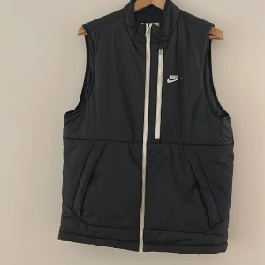 Svart dunväst från Nike Therma-Fit - Svart dunväst från Nike Therma-Fit med vit dragkedja och logga på bröstet. Västen har hög krage, två snedställda fickor fram och är tillverkad i syntetmaterial. Perfekt för lager-på-lager och sportiga outfits.