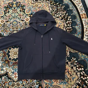 Svart hoodie från Polo Ralph Lauren - Svart hoodie från Polo Ralph Lauren med dragkedja, huva och snörning. Klassisk design med broderad logga på bröstet och två fickor framtill. Perfekt för en chill och stilren look. Den är solbränd i luvan, på längst ut i armarna och midjan men man märker inte till.