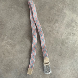  rosa och blått bälte - Snyggt flätat bälte i rosa och blått med beige skinn-detaljer och silverspänne. Bältet har ett coolt mönster och passar perfekt för att ge outfiten lite extra färg. Märkt med 'FASHION' på lädret vid spännet.