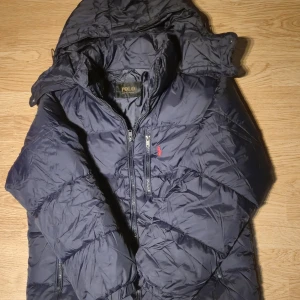 Mörkblå pufferjacka från Polo Ralph Lauren - Säljer en mörkblå pufferjacka från Polo Ralph Lauren med huva och dragkedja framtill. Jackan har en klassisk quiltad look, två sidofickor med dragkedja och Polo-logga broderad i rött på bröstet. Perfekt för kalla dagar och riktigt snygg streetstil.