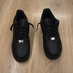 Nike Air Force 1 helsvarta sneakers - Säljer ett par klassiska Nike Air Force 1 i helsvart. Skorna har en låg siluett, svart skinnöverdel, svarta snören och en robust platt sula. Swoosh-loggan är ton-i-ton och de har en silverfärgad detalj vid snörningen. Perfekta för en clean och stilren look. (OPPS ingen kvitto)