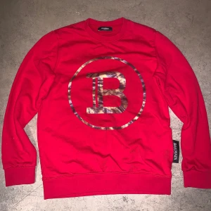Röd sweatshirt Balmain  - Oanvänd helt ny Riktigt snygg röd sweatshirt från Balmain med stort B-logga i metallicmönster på bröstet. Klassisk rund halsringning och långa ärmar. Perfekt statement-plagg för dig som gillar exklusiva streetwear-vibbar. Passar även S, Möts upp i Sthlm