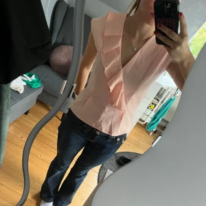 Ljusrosa omlottblus med volang - Supersöt ljusrosa blus i satinliknande material med omlott och volangdetalj framtill. Blusen är ärmlös och har en feminin v-ringning. Perfekt för dig som gillar en romantisk och chic stil. Stängs med dragkedja i ryggen. Omsydd från byxdress till topp av mig❤️XS-S