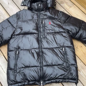 Ralph Lauren Jacka - Svart pufferjacka från Polo Ralph Lauren med glansig finish, röd broderad logga på bröstet och tre dragkedjeförsedda fickor. Jackan har huva, hög krage och långa ärmar. Perfekt för kalla dagar och riktigt snygg streetstil. Storlek M för fler bilder eller frågor. Tveka inte att skriva.