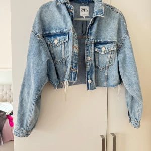 Croppad jeansjacka från Zara - Snygg croppad jeansjacka från Zara i ljusblå denim. Jackan har rå, fransig nederkant, klassisk krage och två bröstfickor med knappar. Perfekt för dig som vill ha en trendig och avslappnad look.